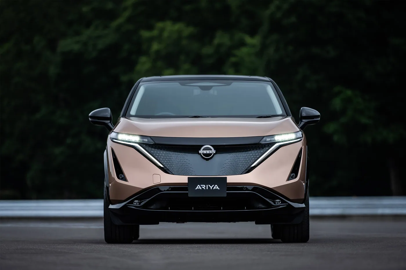 Nissan Ariya cuivrée vue de face, phares allumés, devant une forêt sombre
