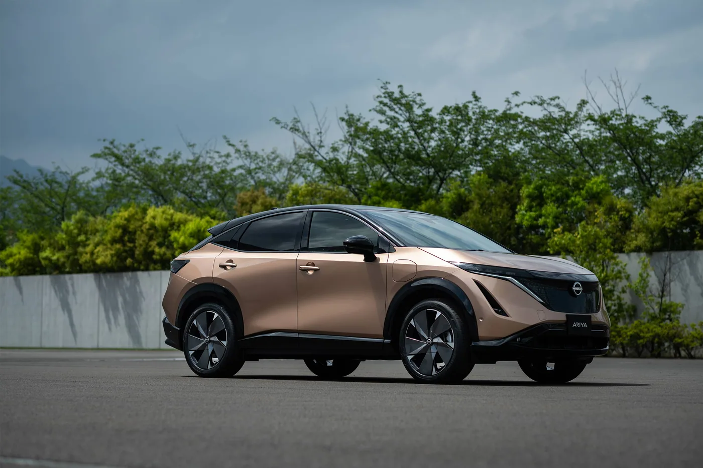 Nissan Ariya cuivrée à toit noir en trois quarts avant, devant des arbres et un mur de béton