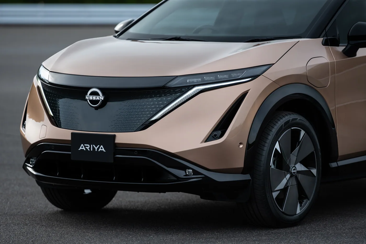 Gros plan sur l'avant d'une Nissan Ariya cuivrée : écusson Nissan, signature lumineuse et plaque Ariya