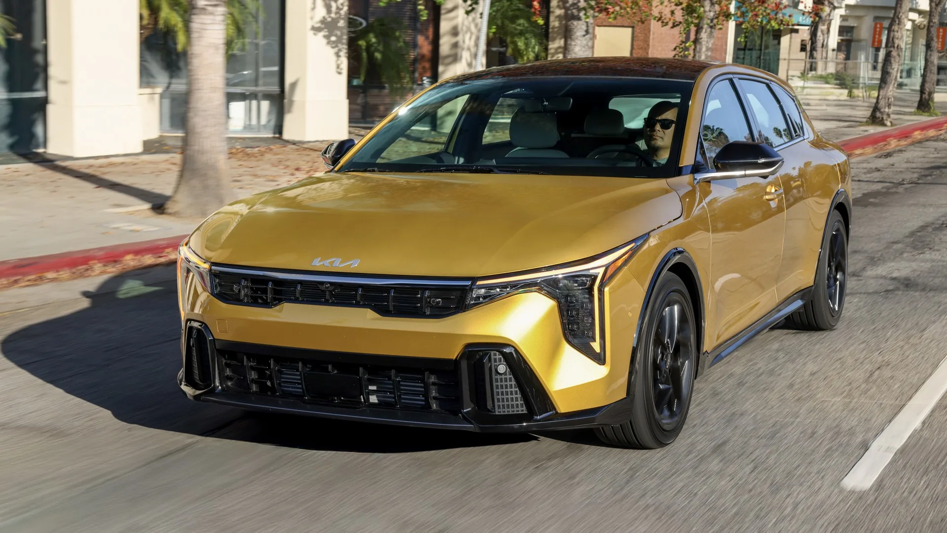 Kia K4 à hayon 2026 jaune roulant dans une rue ensoleillée bordée de commerces