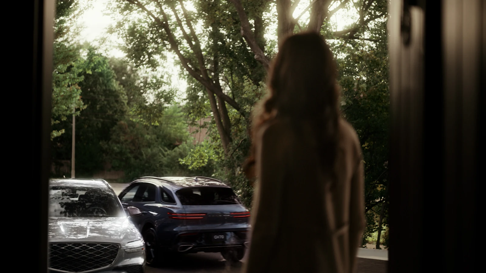 Une femme regarde par sa porte deux Genesis, dont un GV70, stationnées dans son entrée entourée d'arbres