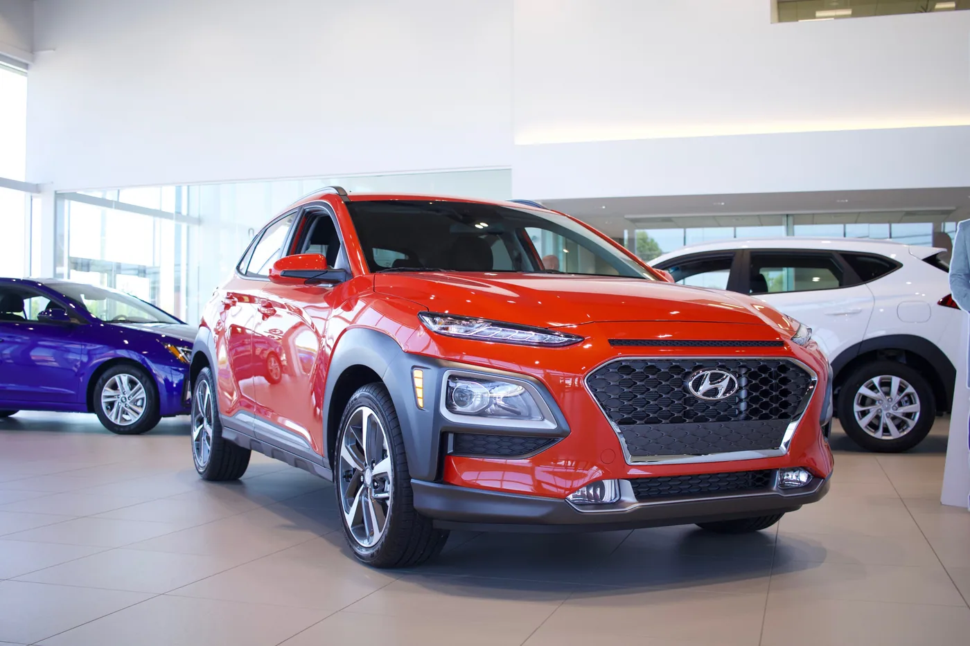 Hyundai Kona orange en trois quarts avant dans une salle d'exposition, entre une berline bleue et un utilitaire blanc