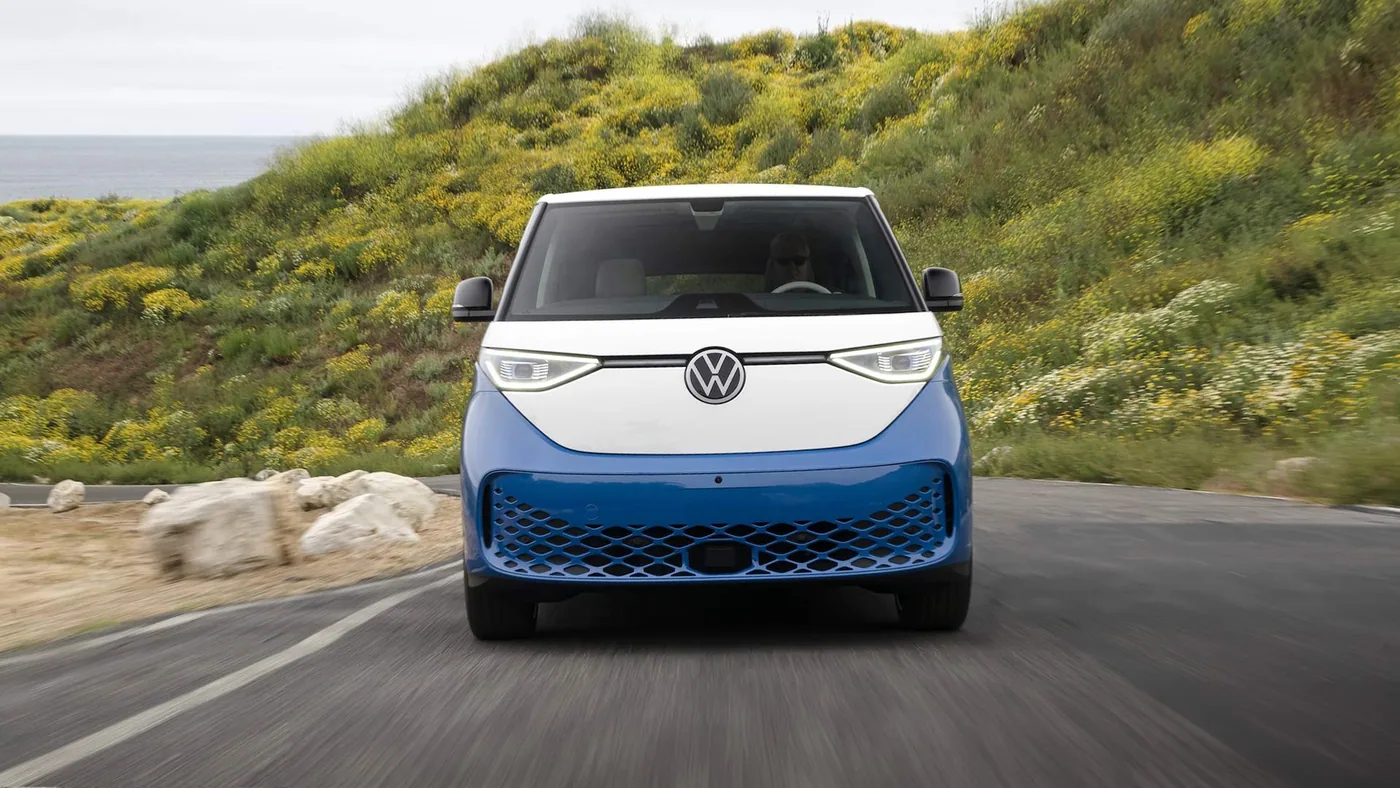 Volkswagen ID. Buzz bleu et blanc vu de face, roulant devant une colline couverte de fleurs jaunes