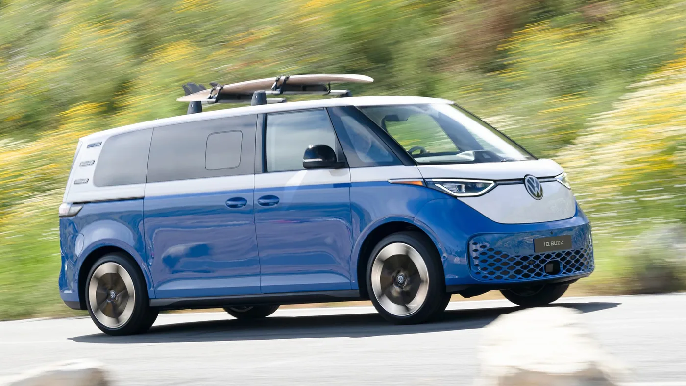 Volkswagen ID. Buzz bleu et blanc en profil, avec une planche de surf sur le toit, roulant sur une route fleurie