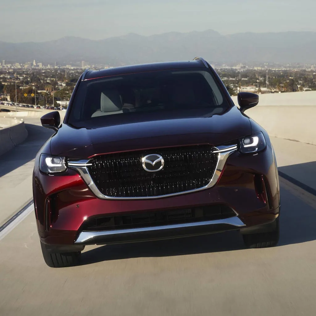 Mazda CX-90 2024 rouge foncé vu de face sur une route en hauteur, ville au loin