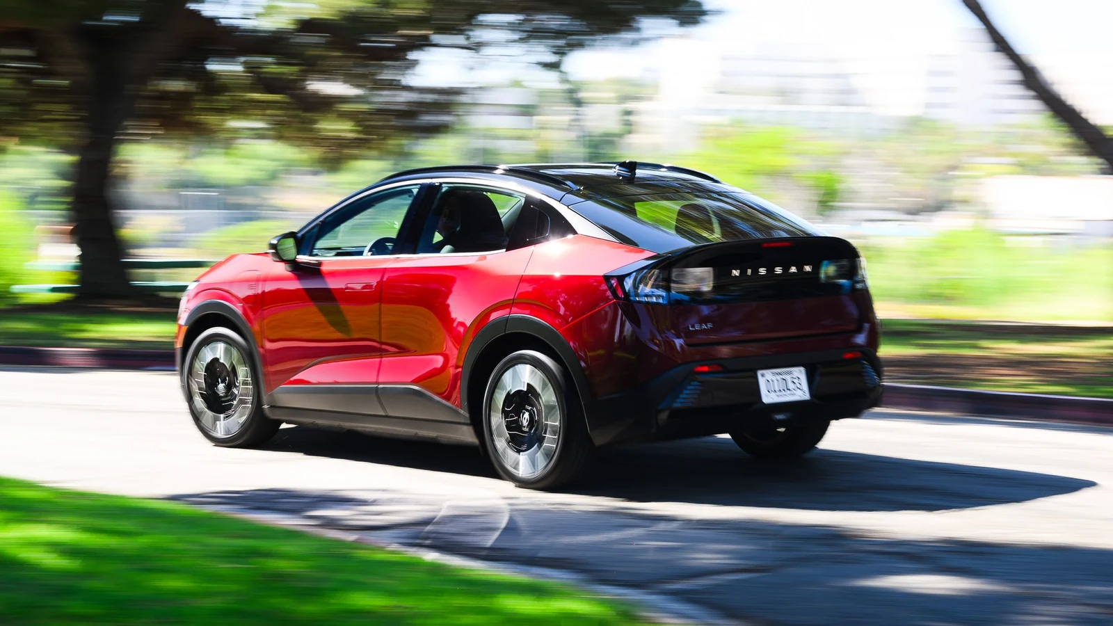 Nissan LEAF rouge à toit noir vue de trois quarts arrière, roulant sur une route bordée d'arbres et de gazon