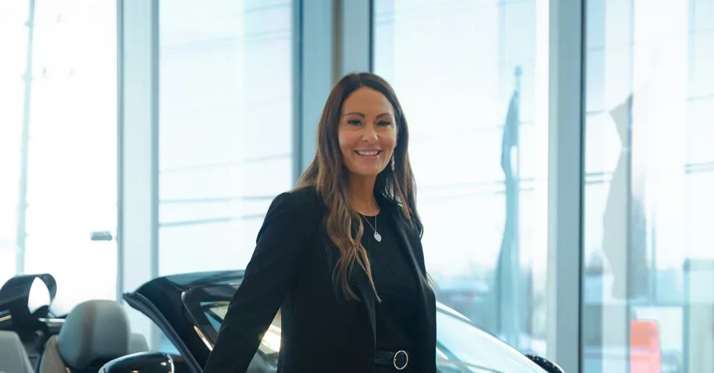 Mariève Beaucage souriante dans une salle de montre lumineuse, véhicule décapotable exposé derrière elle