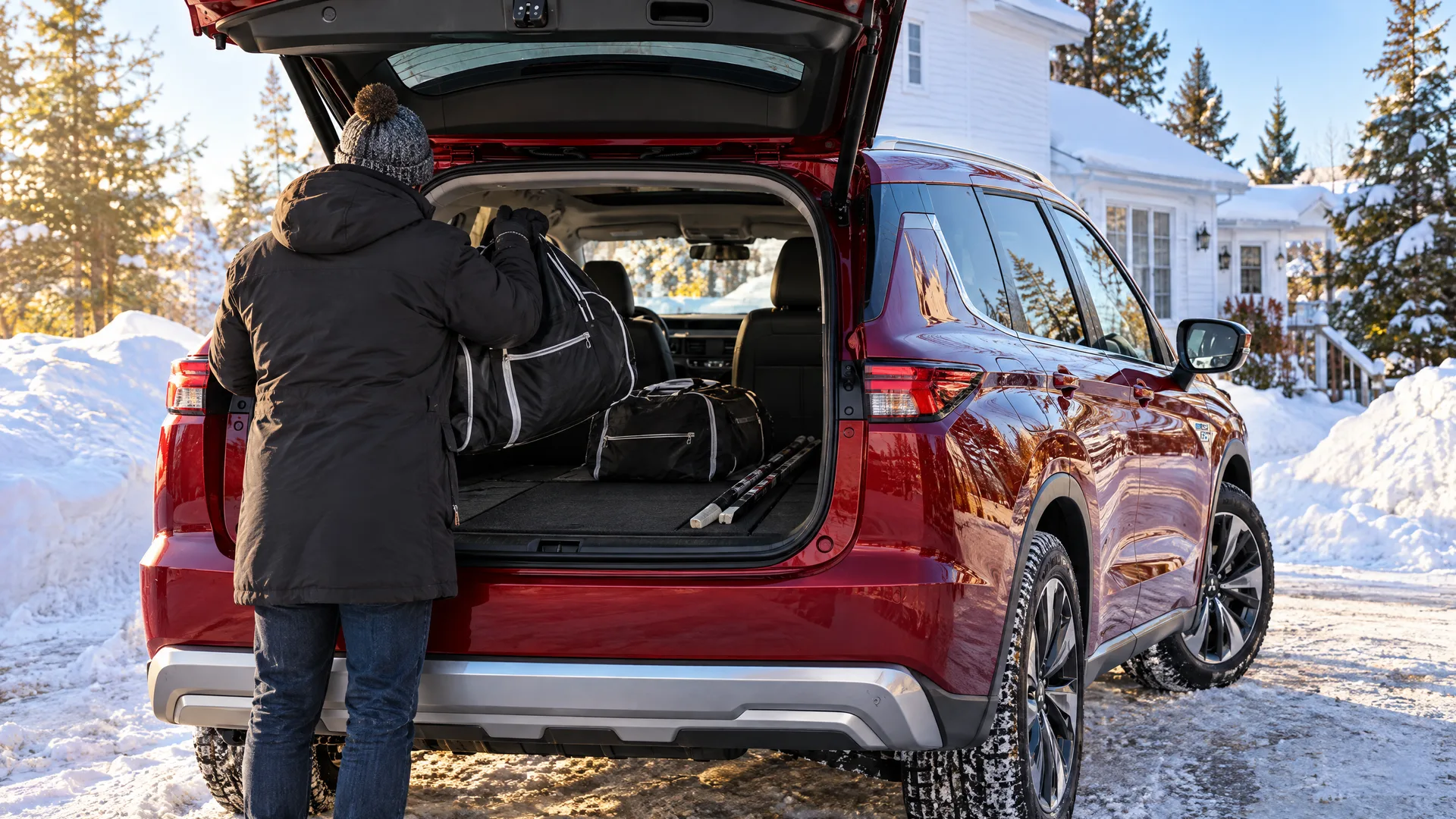 Personne de dos qui charge des sacs d'équipement de hockey dans le coffre d'un Mitsubishi Outlander PHEV 2026, troisième rangée rabattue, entrée résidentielle en hiver