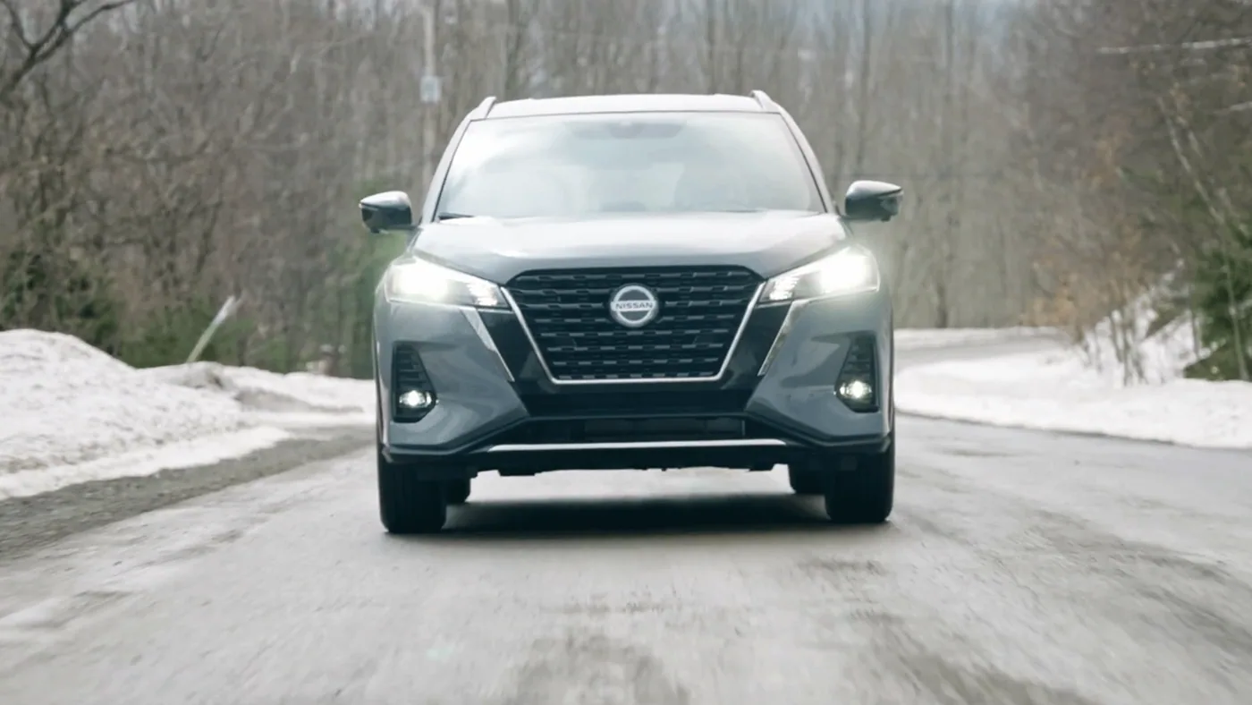 Nissan Kicks 2021 gris vu de face, phares allumés, roulant sur une route de campagne enneigée