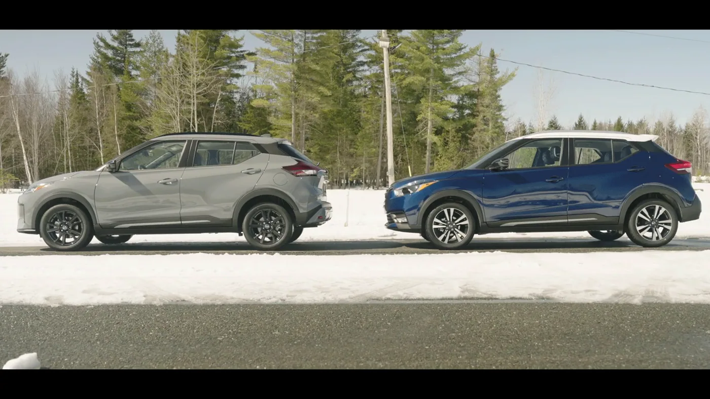 Nissan Kicks 2021 gris et ancien Kicks bleu à toit blanc vus de profil, côte à côte, devant des sapins enneigés
