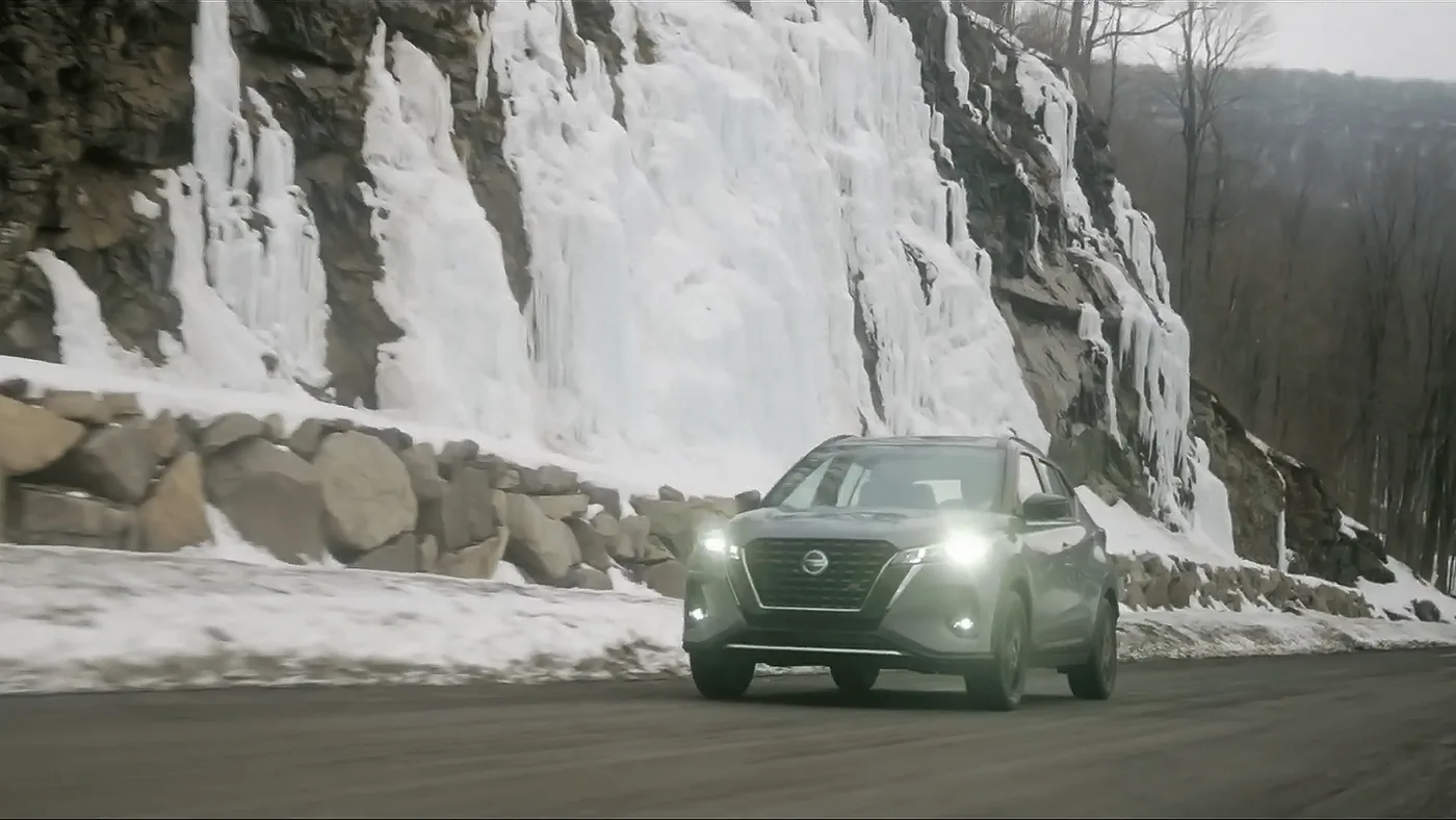Nissan Kicks 2021 gris roulant phares allumés devant une paroi rocheuse couverte de glace, en hiver