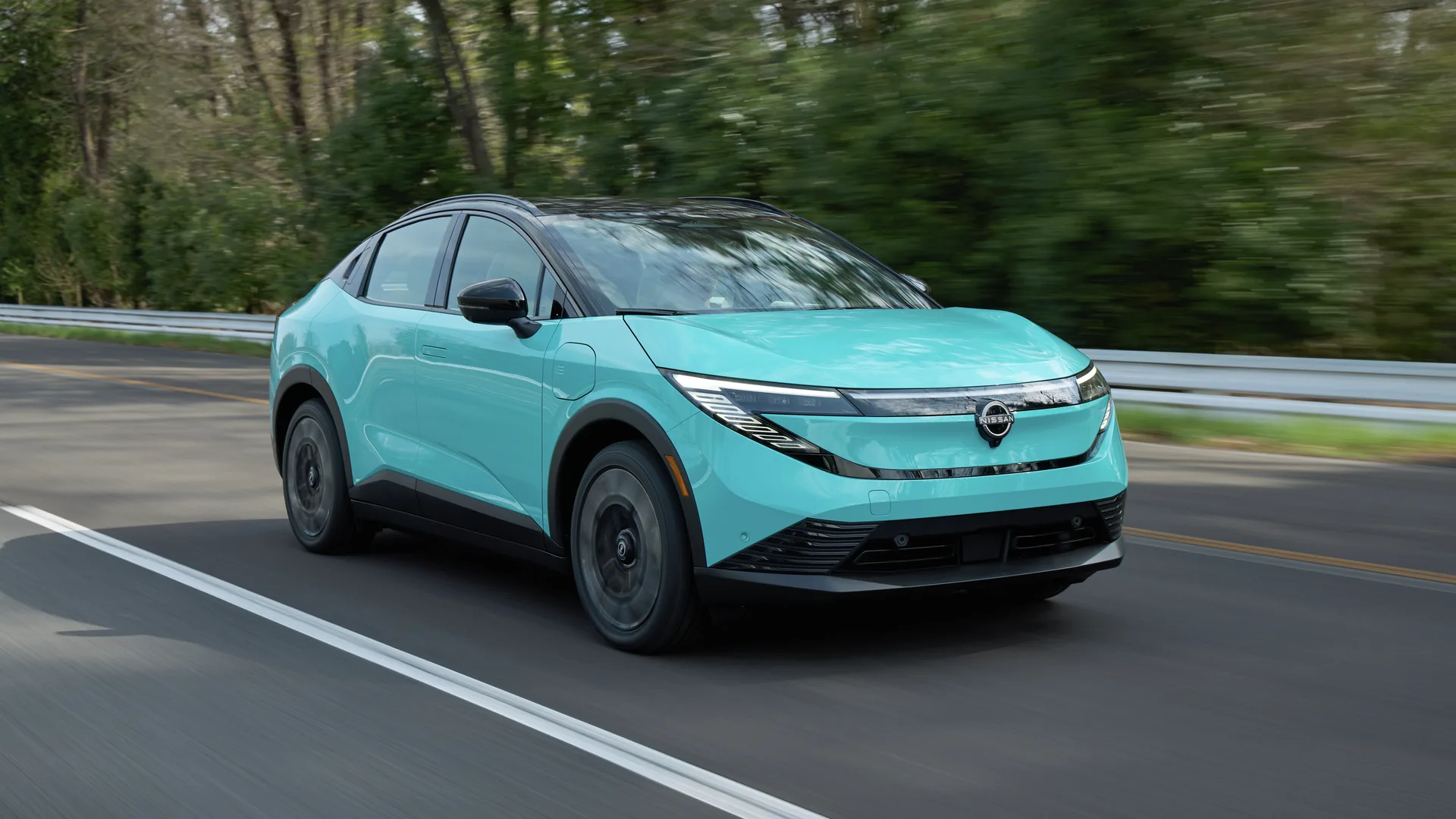 Nissan LEAF turquoise vue de trois quarts avant, en mouvement sur une route boisée bordée d'un garde-fou