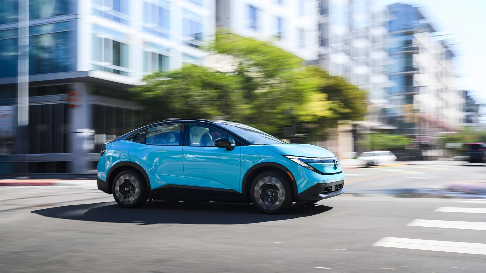 Nissan LEAF bleu clair roulant sur un boulevard urbain bordé d'immeubles, vue de trois quarts avant