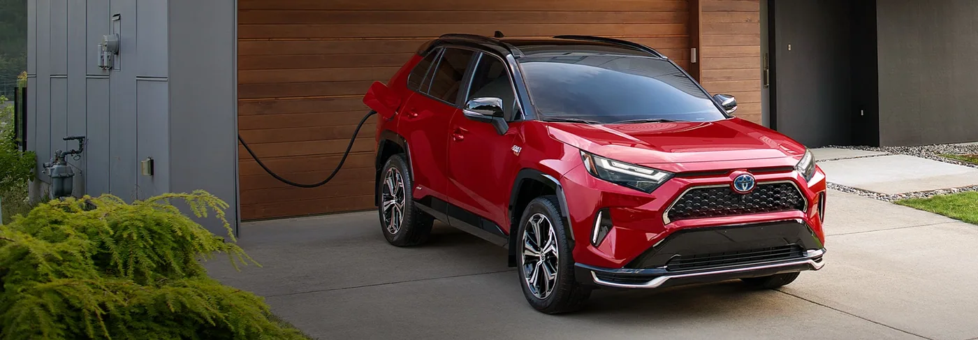 Toyota RAV4 Prime rouge branché à une borne de recharge devant un garage, vue de trois quarts avant