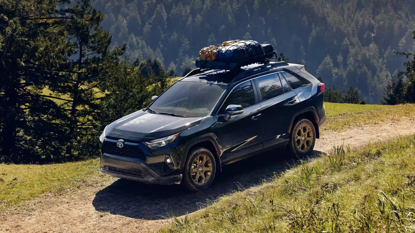 Toyota RAV4 2023 noir avec bagages sur le toit, sur un sentier de montagne bordé de conifères