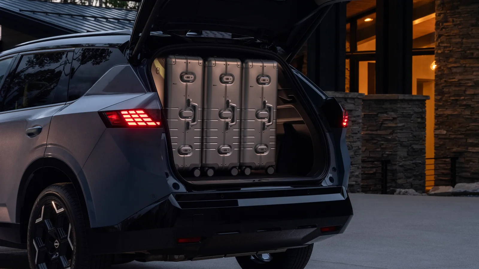 Hayon ouvert d'un Nissan Rogue hybride 2027 chargé de trois grandes valises rigides, devant une maison au crépuscule