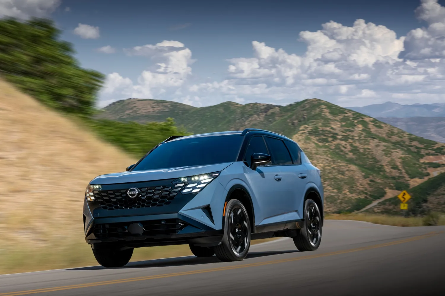 Nissan Rogue hybride 2027 bleu en mouvement sur une route de montagne, vu de trois quarts avant