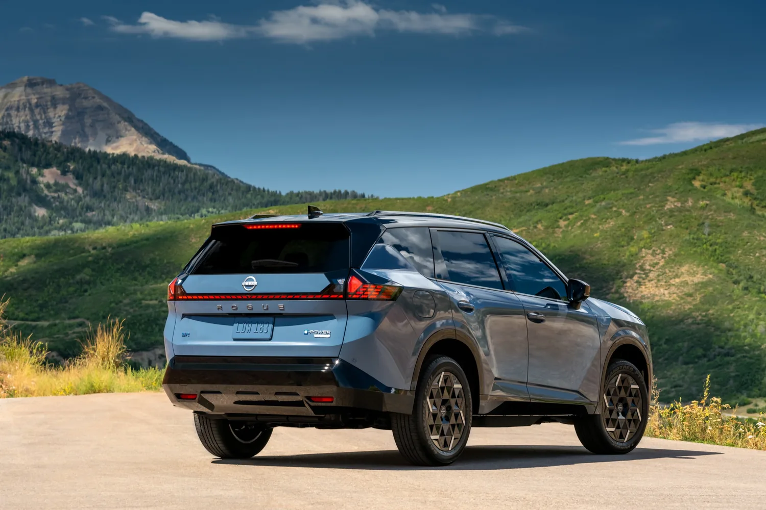 Vue arrière d'un Nissan Rogue hybride 2027 bleu sur une route de montagne, feux en bandeau et lunette teintée