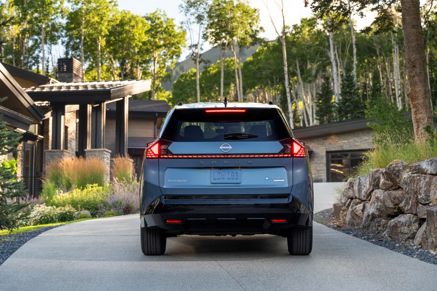 Nissan Rogue hybride 2027 bleu vu de l'arrière dans l'allée d'une maison entourée de bouleaux
