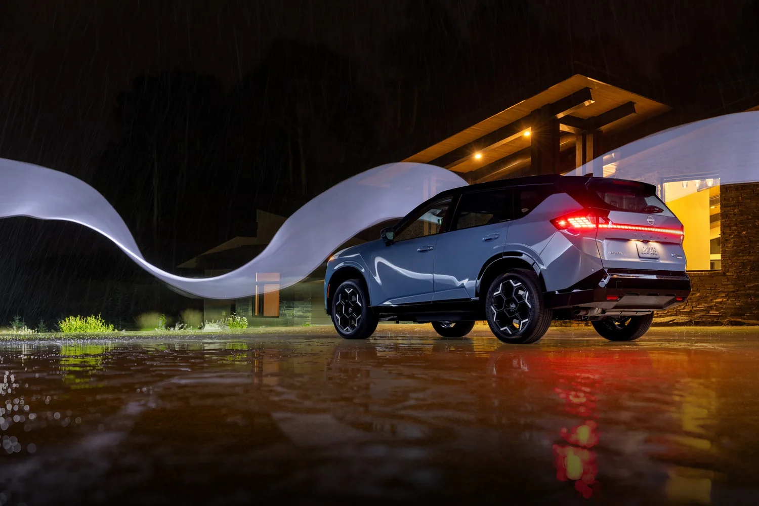 Nissan Rogue hybride 2027 bleu de nuit sous la pluie devant une maison éclairée, reflets sur l'asphalte mouillé