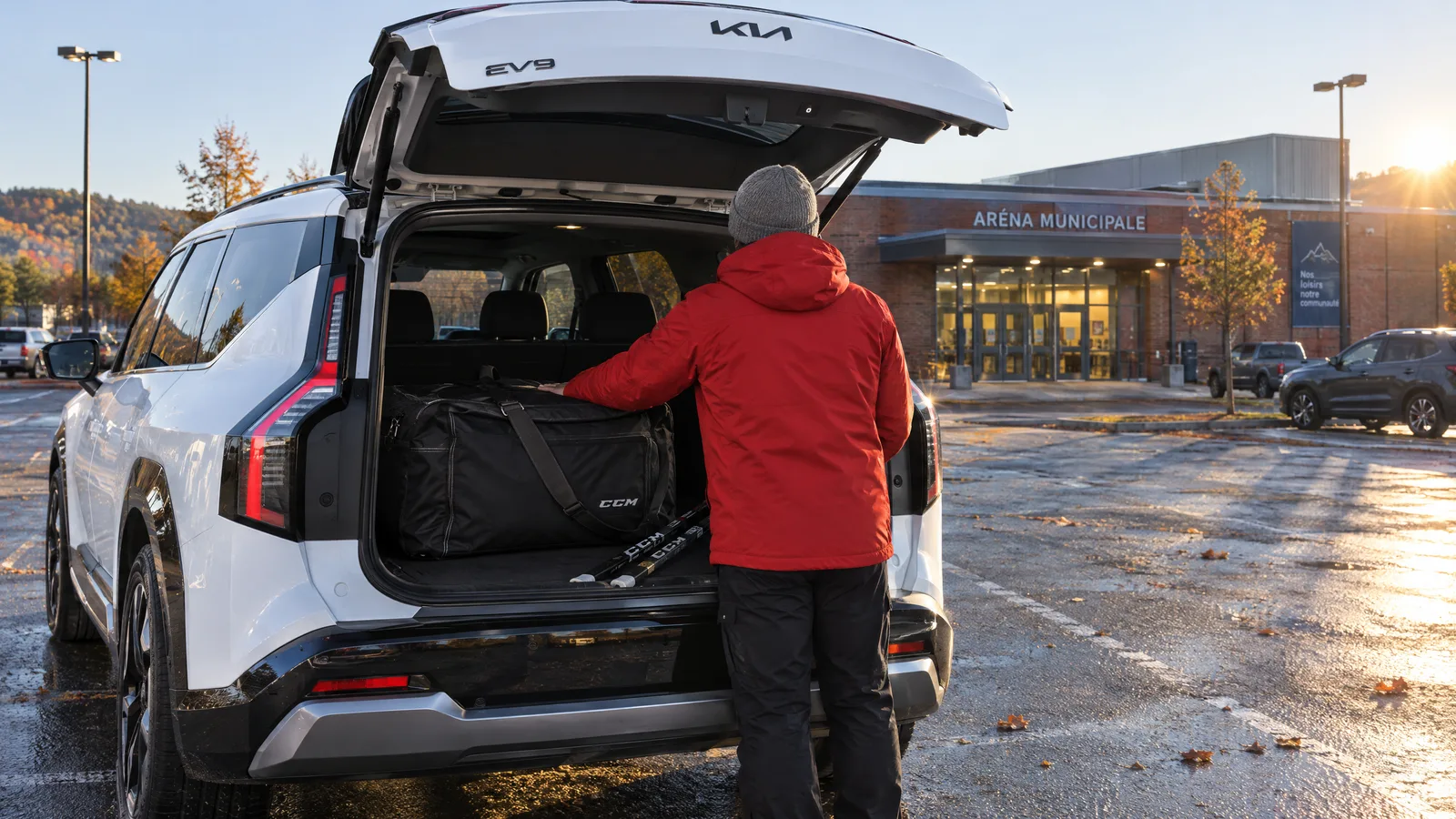Personne de dos qui glisse un sac de hockey dans le coffre ouvert d'un Kia EV9 blanc, stationnement d'aréna en fin de journée d'automne