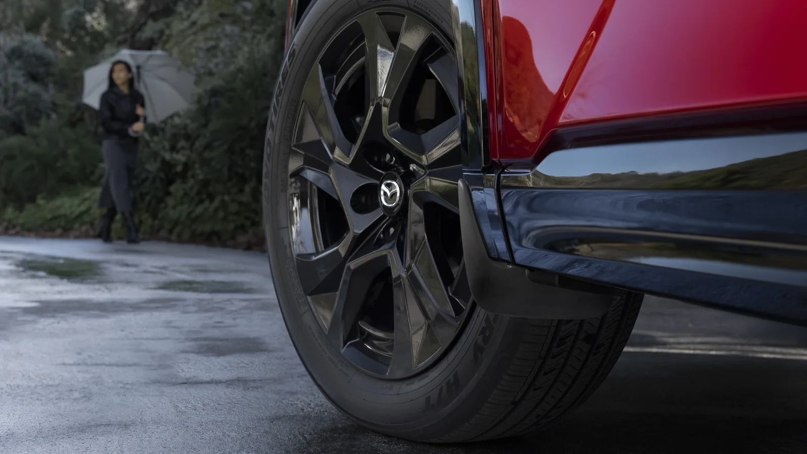 Gros plan de la roue avant d'un Mazda CX-5 rouge sur une chaussée mouillée, une passante avec un parapluie en arrière-plan