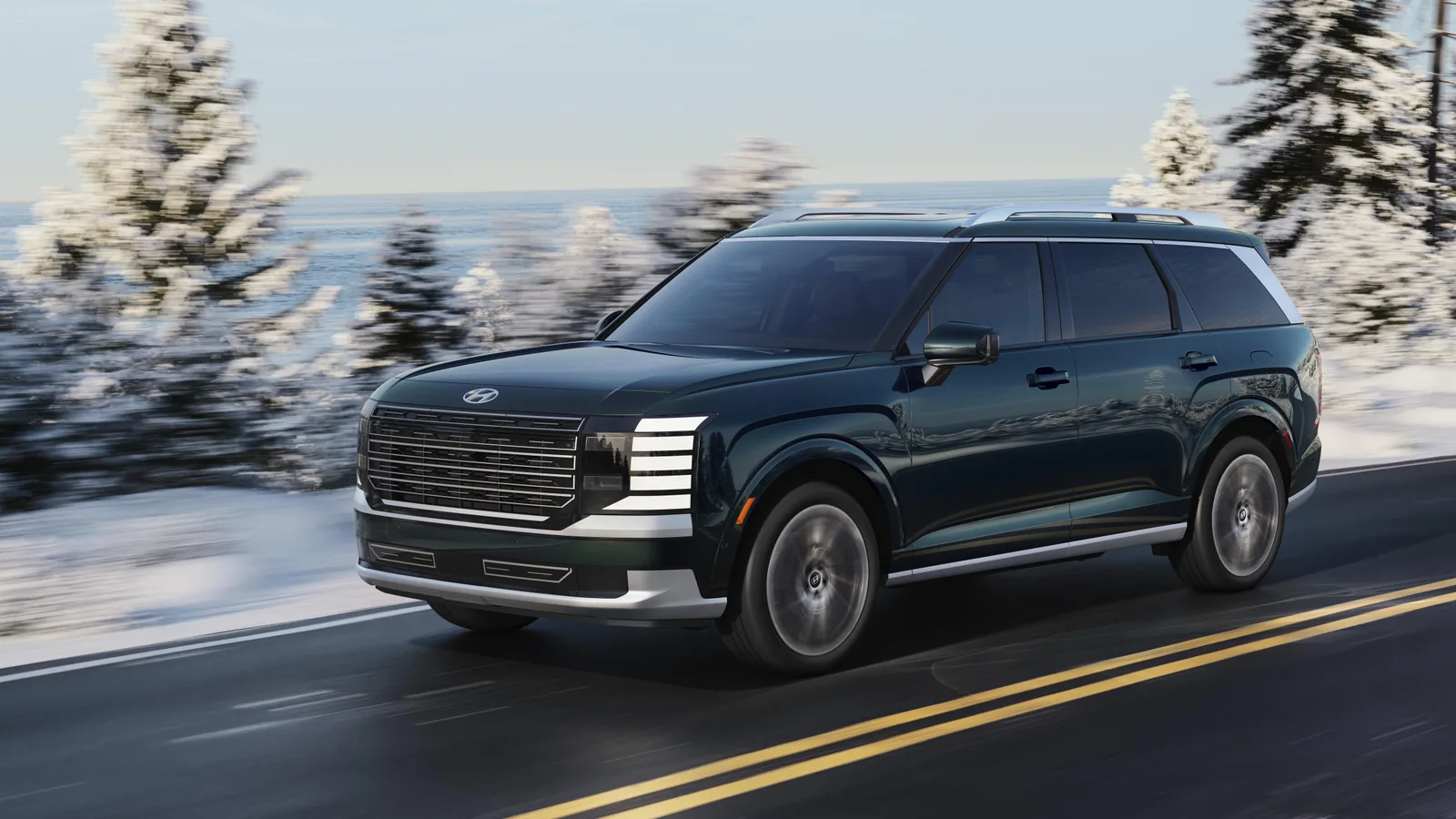 Hyundai Palisade vert foncé roulant sur une route dégagée bordée de neige et de sapins givrés