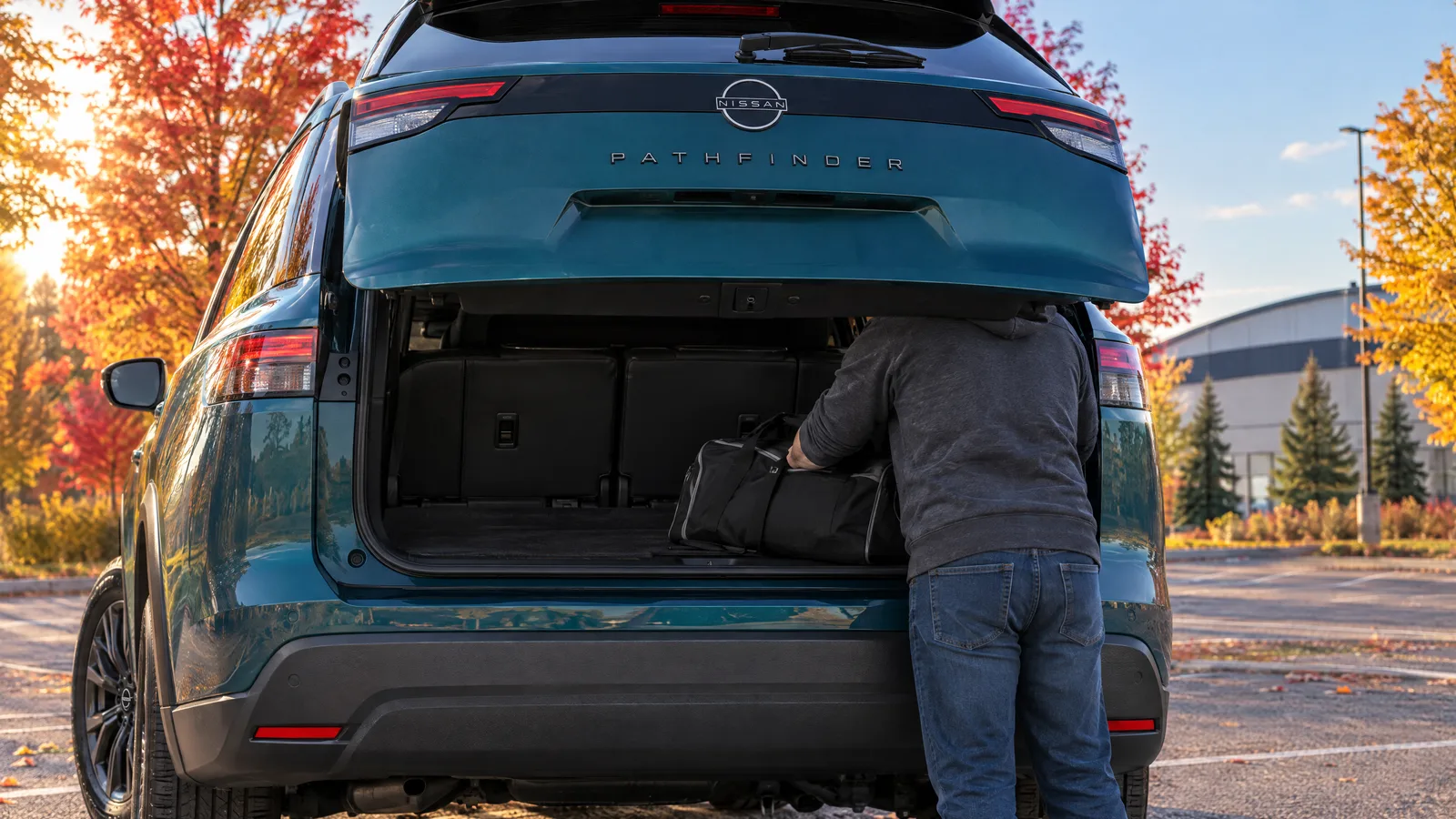Personne de dos qui installe un sac de sport dans le coffre d'un Nissan Pathfinder 2026 sarcelle, stationnement d'aréna en fin de journée d'automne