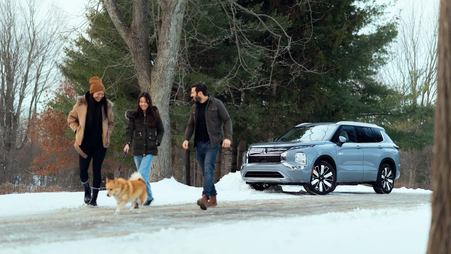 Famille et son chien marchant dans la neige à côté d'un Mitsubishi Outlander PHEV 2026 bleu pâle