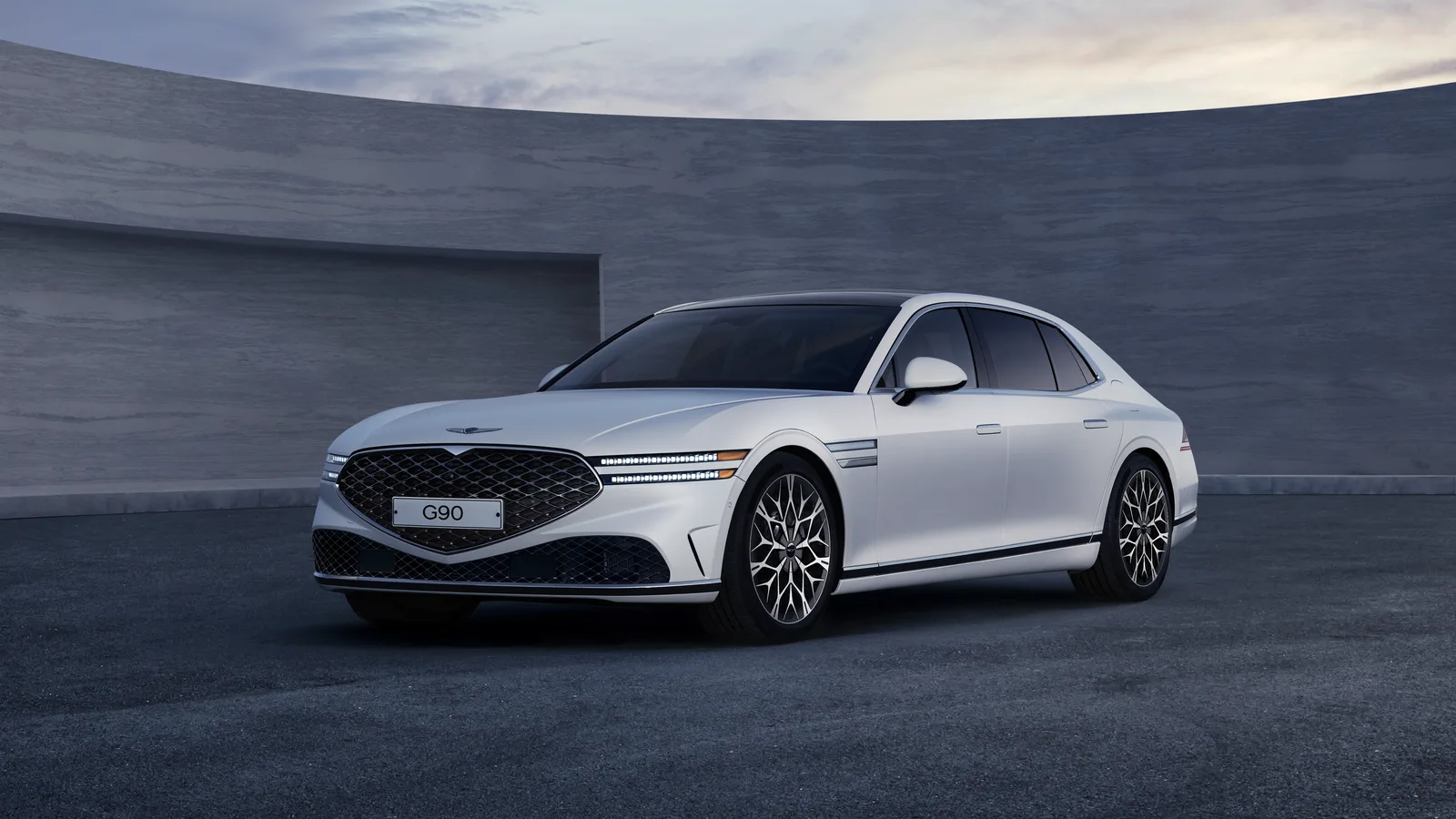 Genesis G90 blanche entière, vue de trois quarts avant devant des murs courbes en béton au crépuscule.
