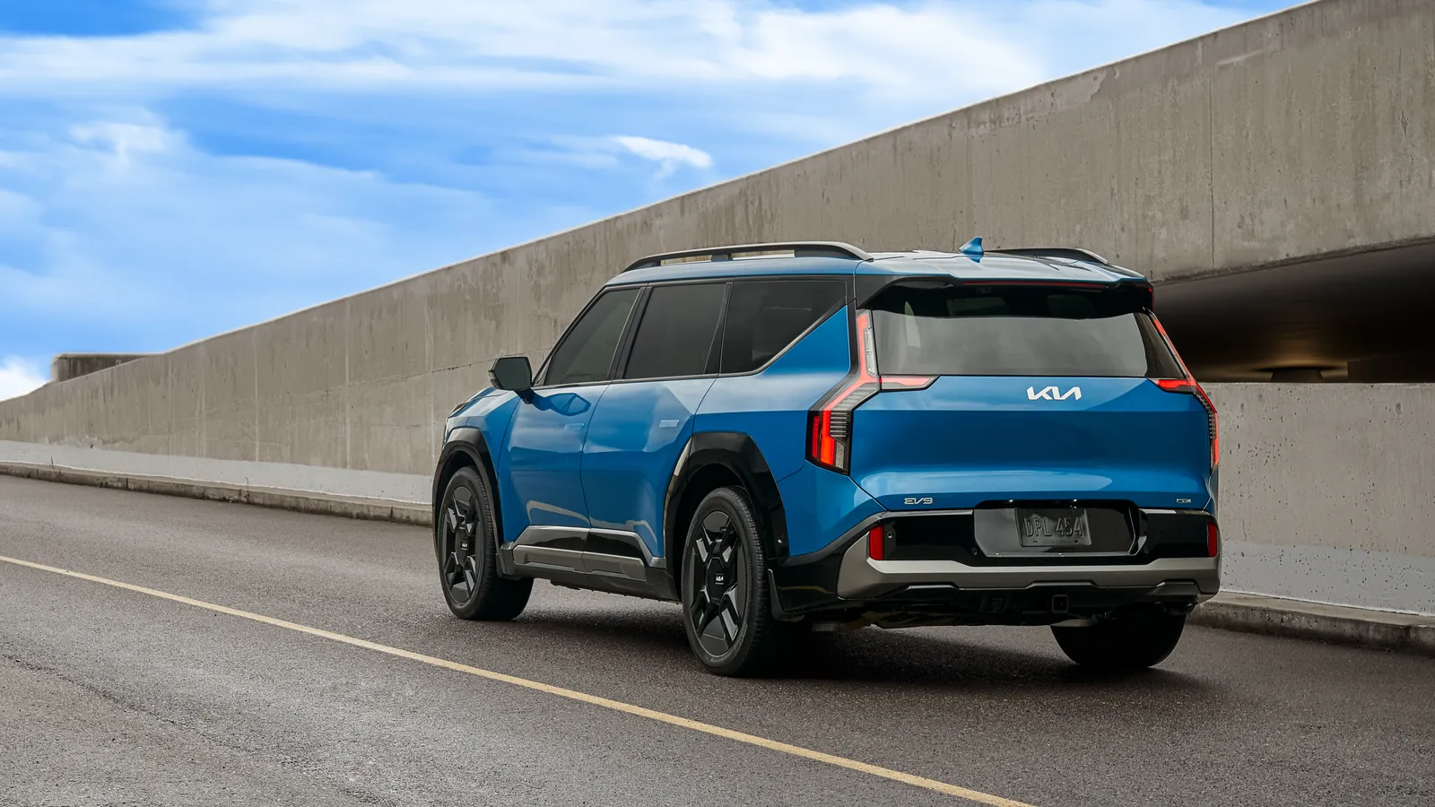 Kia EV9 2024 bleue entière vue de trois quarts arrière sur une route bordée d’un mur de béton.