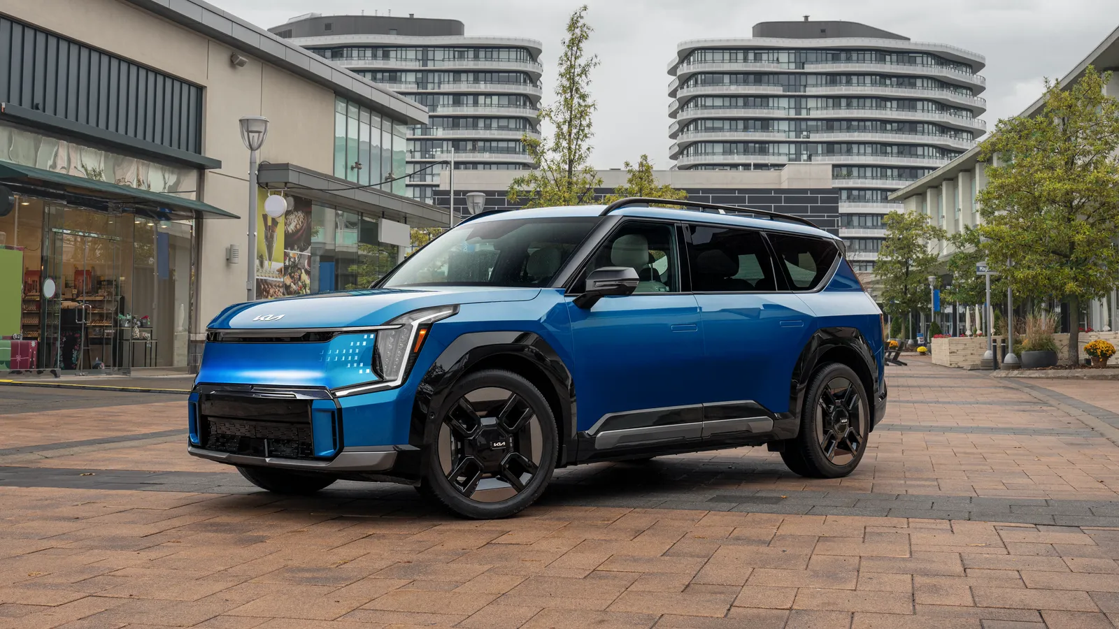 Kia EV9 2024 bleue entière, vue de trois quarts avant dans une rue bordée d’immeubles contemporains.