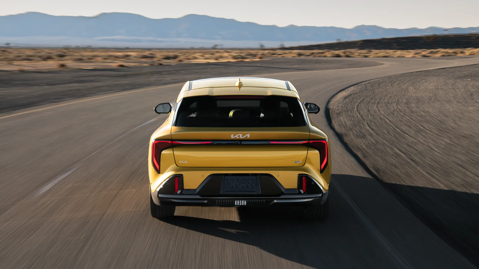 Kia K4 à hayon 2026 jaune vue de l’arrière dans un virage, logo Kia et feux verticaux visibles.