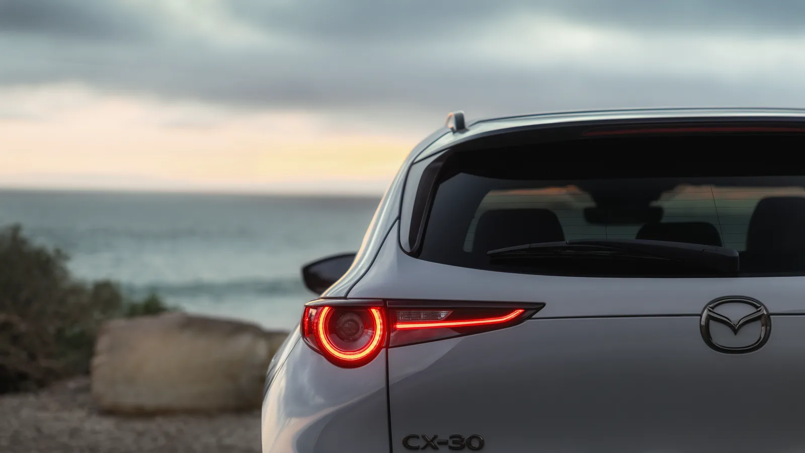 Emblème Mazda et feu arrière circulaire d’un CX-30 2026 argent, avec la mer à l’arrière-plan.