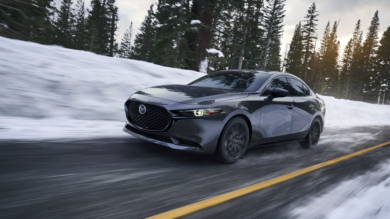 Mazda3 2026 grise entière, vue de trois quarts avant sur une route enneigée bordée de conifères.