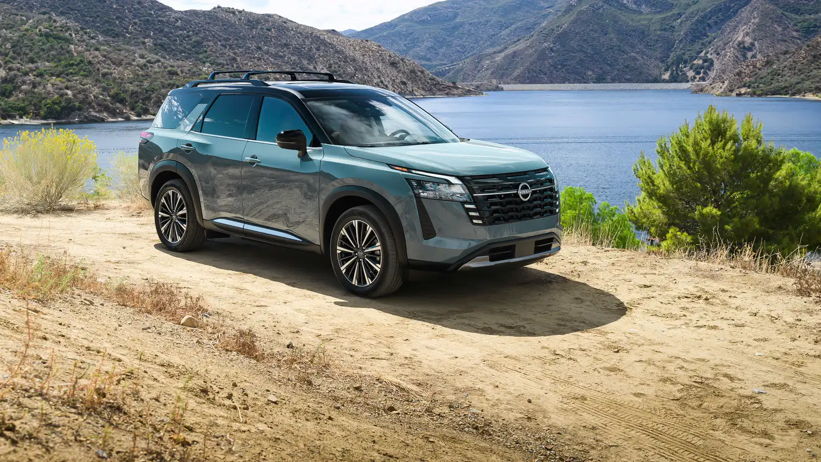 Nissan Pathfinder 2026 bleu-vert entier, vu de trois quarts avant au bord d’un lac, montagnes en arrière-plan.