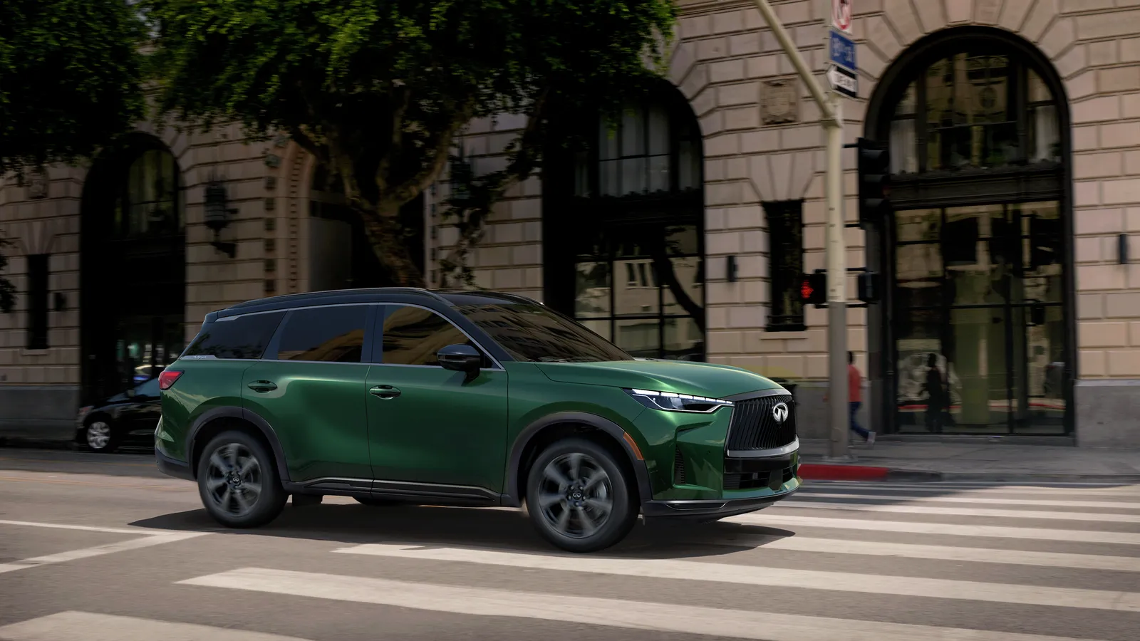 INFINITI QX60 2027 vert en roulage dans une rue urbaine bordée de bâtiments en pierre, piétons hors foyer à l’arrière-plan.