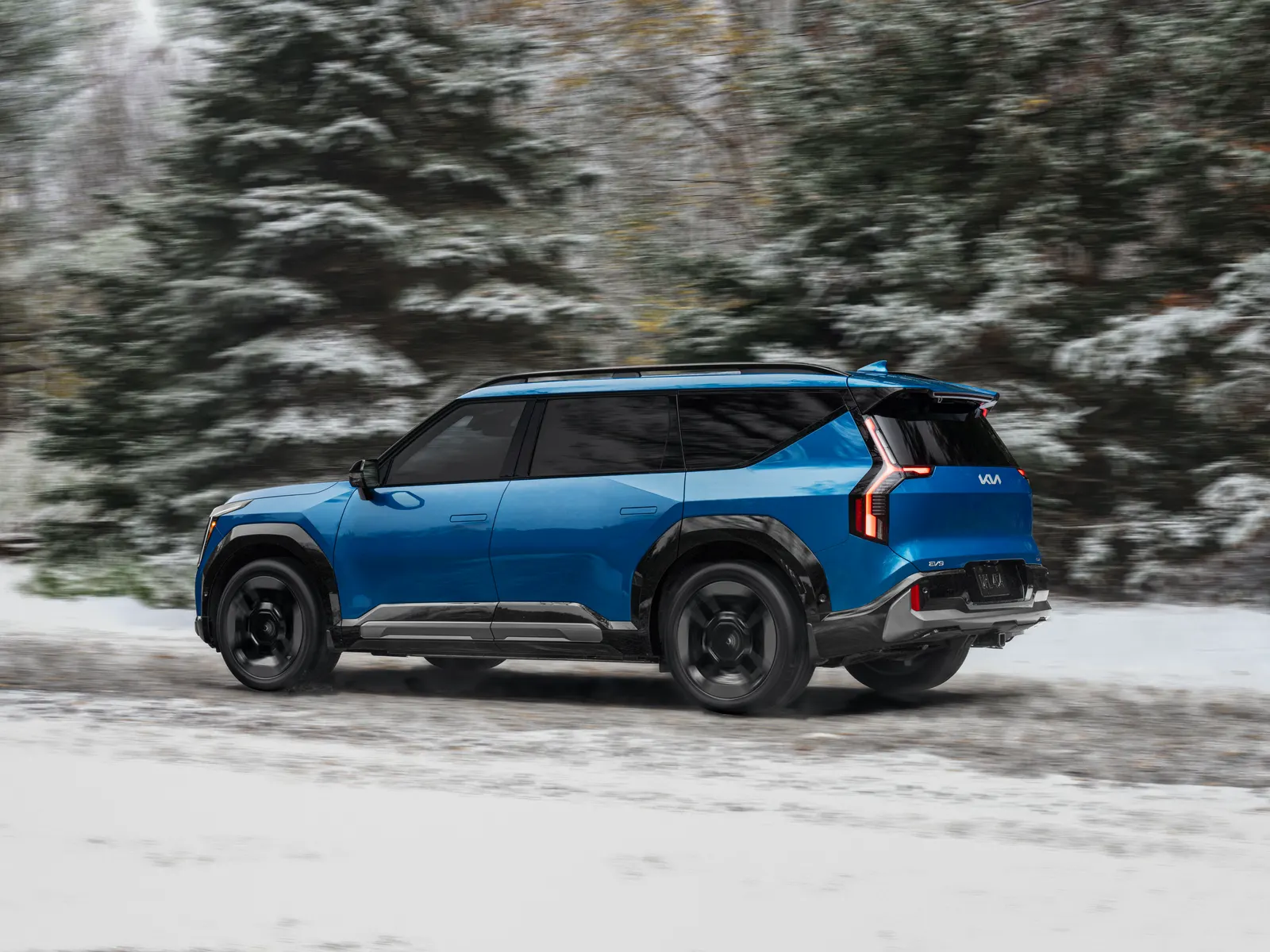 Kia EV9 bleu roulant sur une route enneigée bordée de sapins