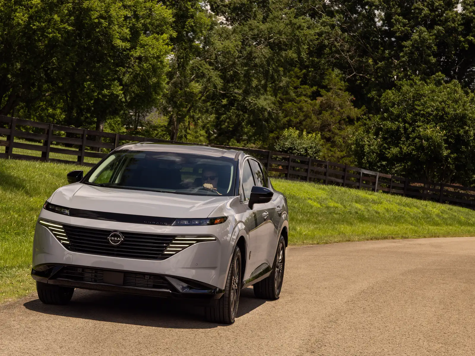 Nissan Murano gris vu de trois quarts avant sur une allée de campagne bordée d’une clôture