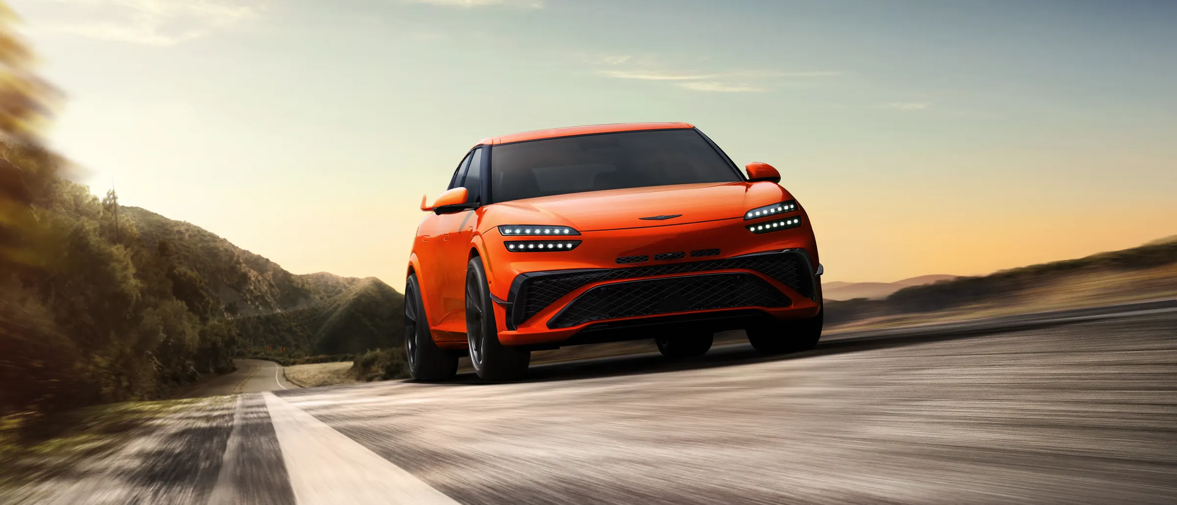 Genesis GV60 Magma orange, VUS électrique, de face en mouvement sur une route de campagne au coucher du soleil.
