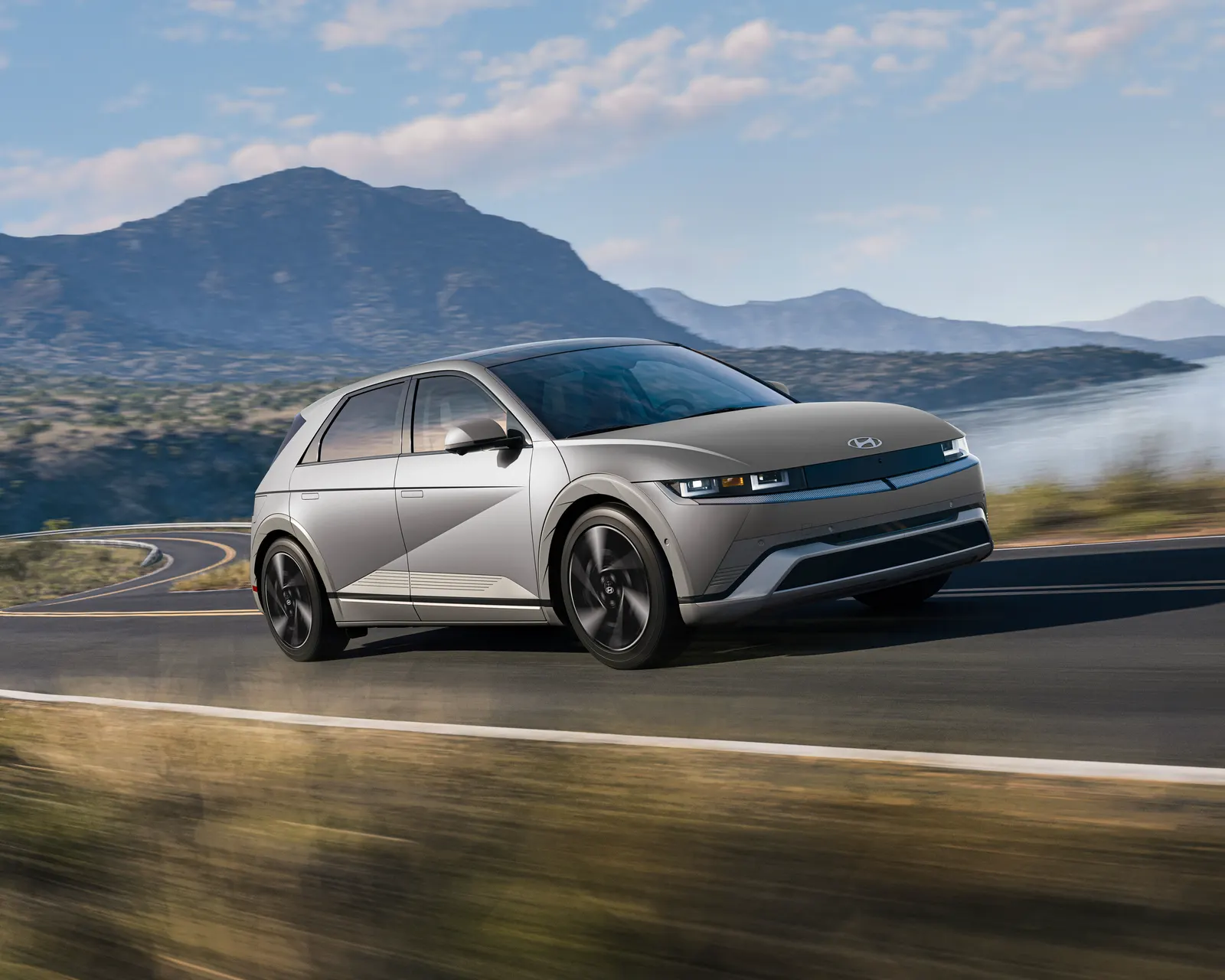 Hyundai IONIQ 5 argent, véhicule électrique, en mouvement sur une route côtière, montagnes et lac en arrière-plan.