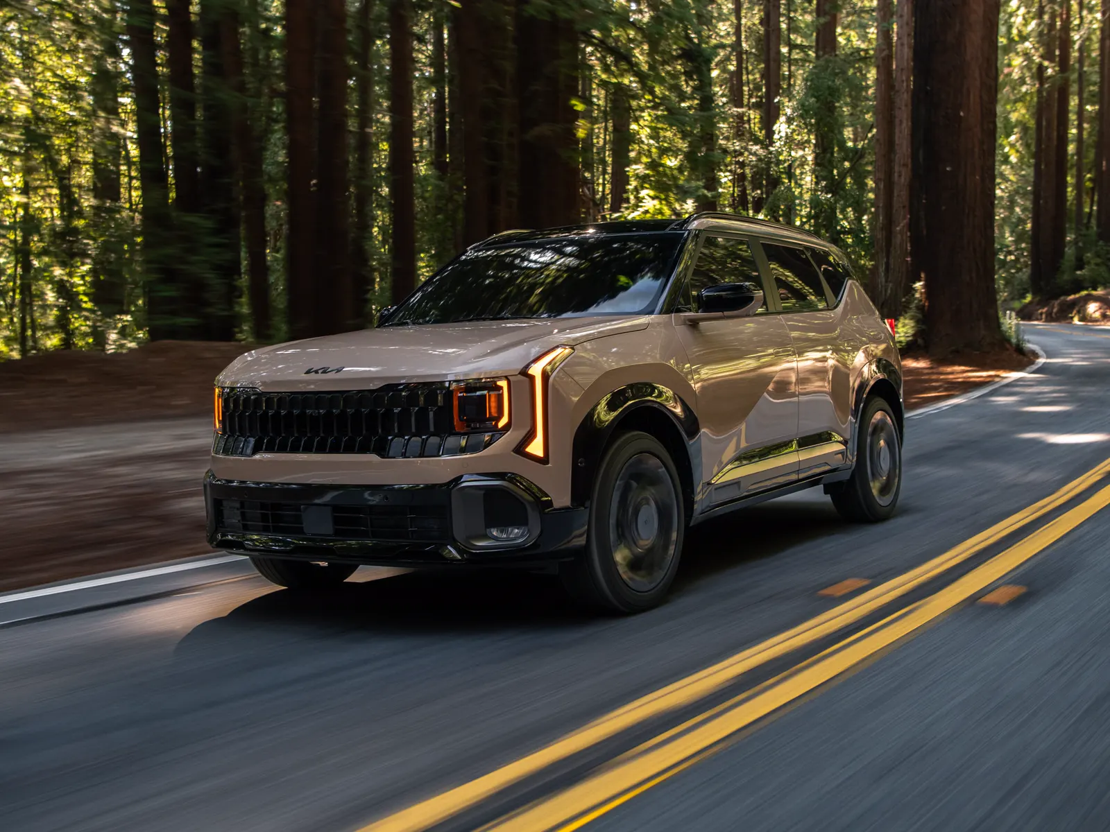 Kia Seltos 2027 beige vu de trois quarts avant sur une route sinueuse en forêt.