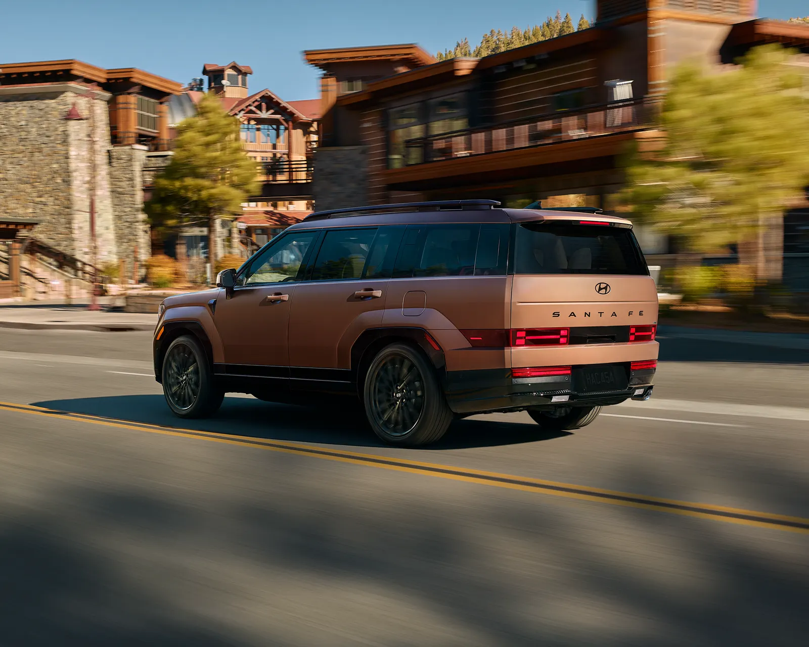 Hyundai Santa Fe 2026 cuivré vu de trois quarts arrière en mouvement dans la rue d'un village de montagne.