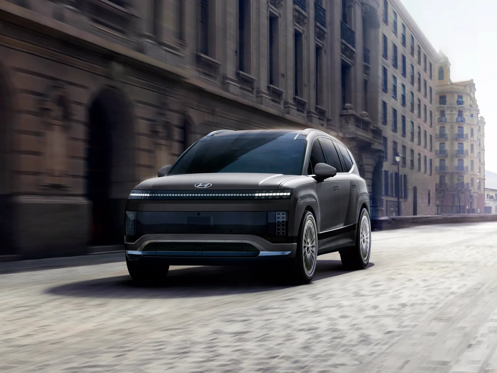 Hyundai IONIQ 9 gris foncé vu de trois quarts avant sur une rue pavée entre de grands immeubles de pierre.