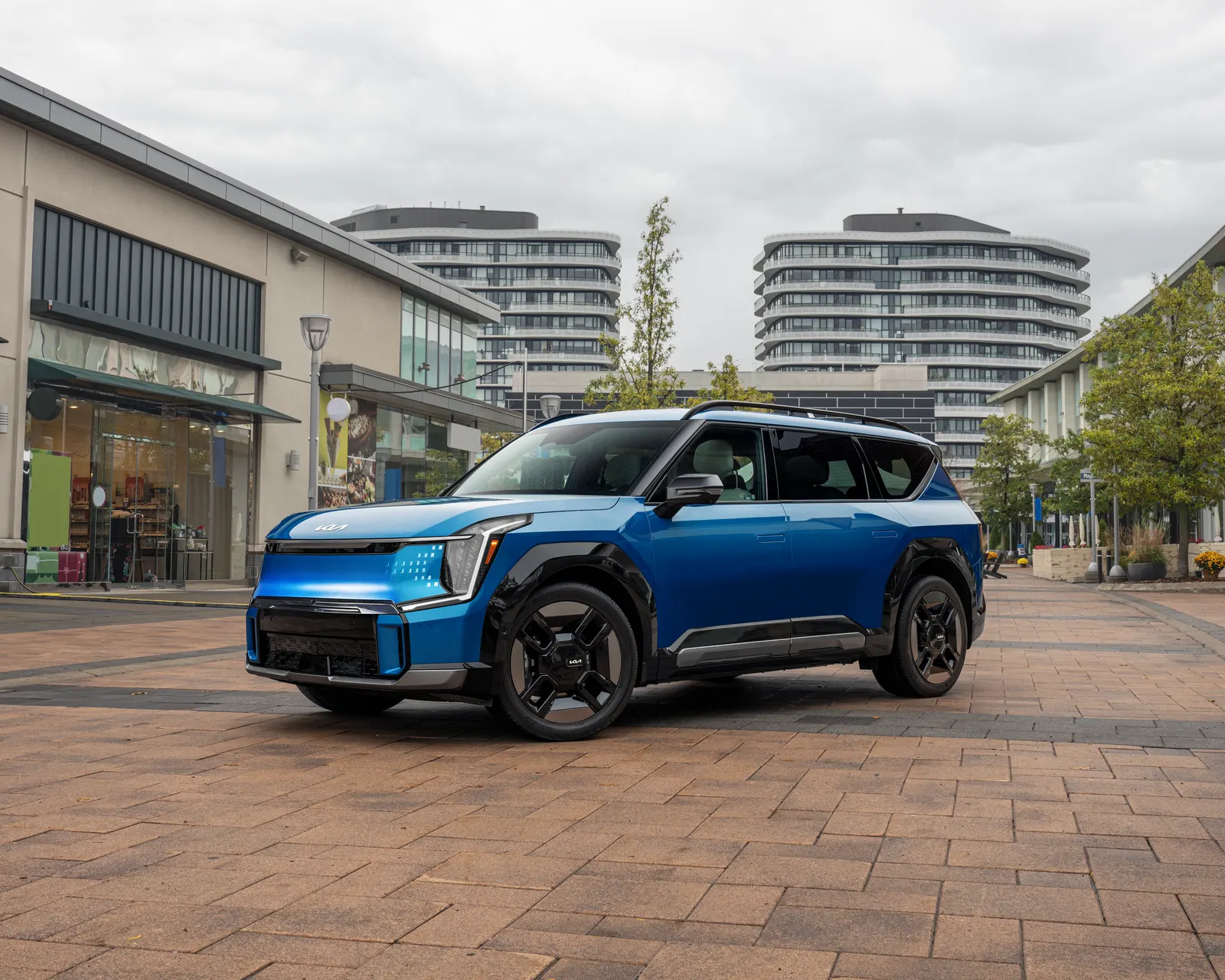 Kia EV9 bleu, VUS électrique à trois rangées, vu de trois quarts avant sur une place piétonne bordée de boutiques.